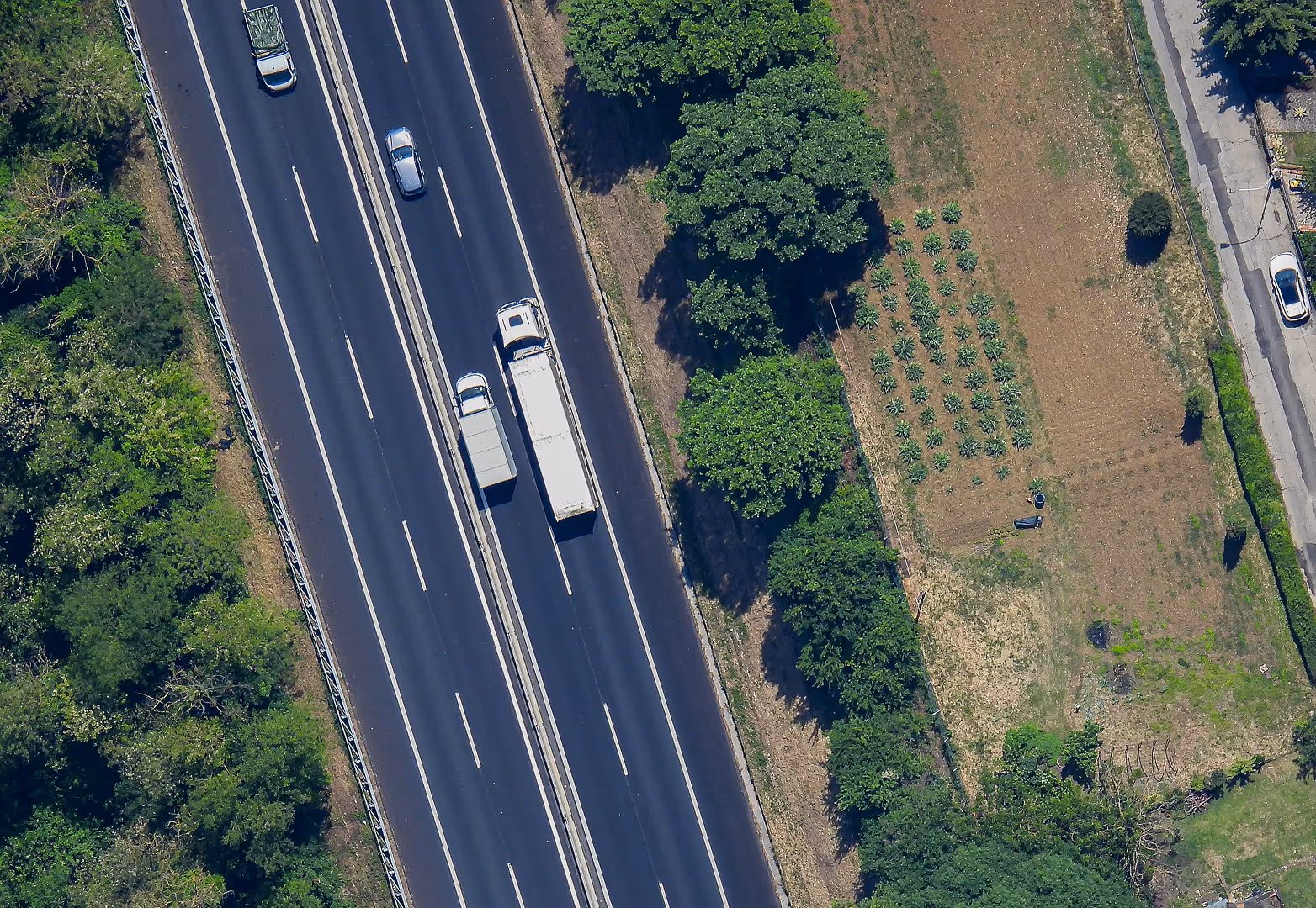 Aerial view of a highway with moving vehicles next to a small cultivated field and tree-lined edge, captured from a Stratostats® high-altitude platform. Cars and trucks are traveling in both directions.