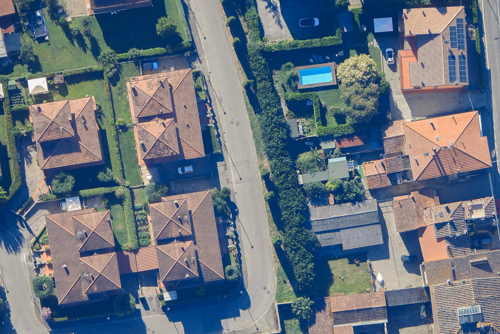 Aerial view of a suburban neighborhood with red-tiled roofs, green lawns, and parked cars, captured from a Stratostats® high-altitude platform. A central road and a backyard pool are visible.